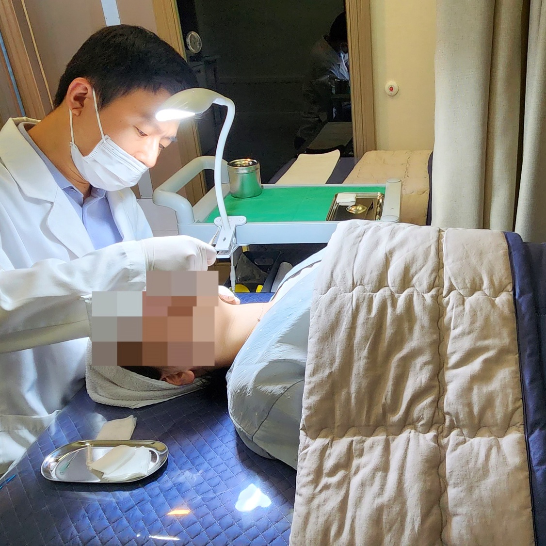마스크를 쓴 원장이 스탠드 조명 아래 누운 환자의 얼굴을 살피며 진행하는 비립종 시술 장면